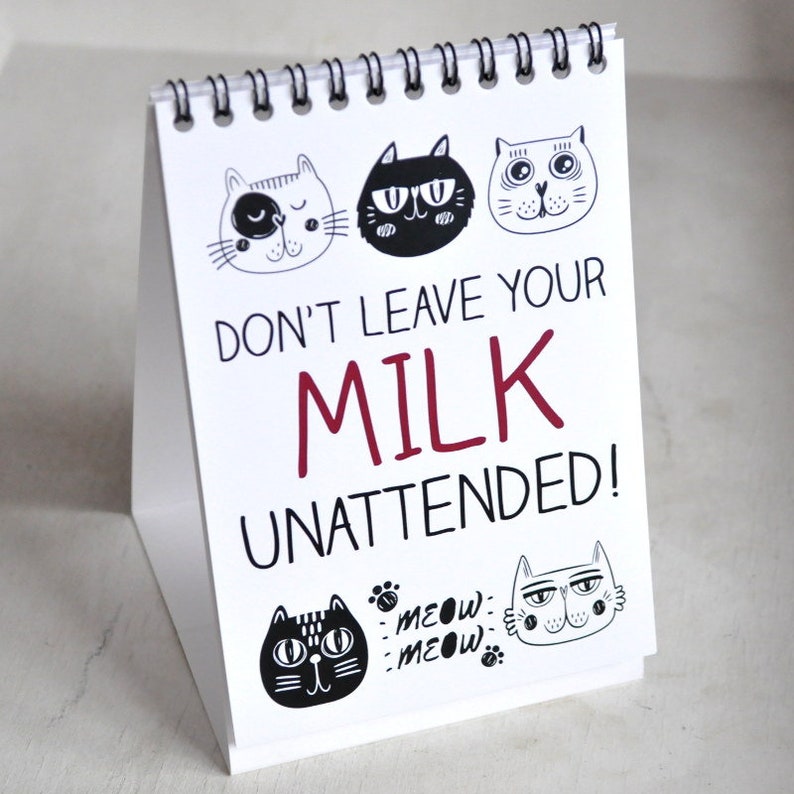 May include: White sign with black and white cat illustrations and the text "Don't leave your milk unattended!" in red and black text. The sign has a spiral binding and is standing upright.