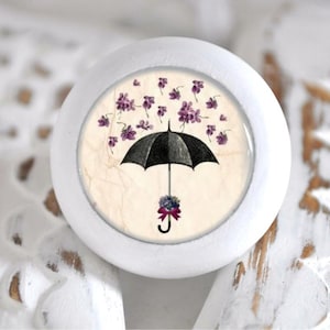 Könnte beinhalten: Weißer Keramik-Knopf mit einem runden, bedruckten Design, das einen schwarzen Regenschirm mit herabfallenden lila Blumen zeigt.