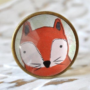 Könnte beinhalten: Ein runder, bronzefarbener Knopf mit einer Fuchsgesichts-Illustration. Der Fuchs hat orangefarbenes Fell, eine weiße Schnauze und schwarze Augen und Nase.