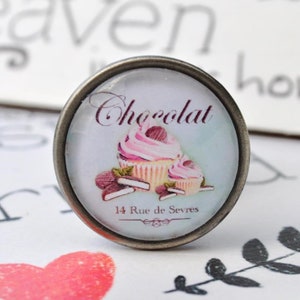 Könnte beinhalten: Ein runder silberfarbener Knopf mit einem Glaseinsatz, der eine Vintage-Illustration eines rosa und weißen Cupcakes mit Schokoladenstücken und dem Text "Chocolat 14 Rue de Sevres" zeigt.