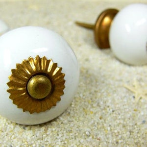 Könnte beinhalten: Zwei weiße Keramikknöpfe mit goldenen Akzenten. Die Knöpfe haben ein dekoratives, sonnenstrahlenartiges Design mit einer goldenen Mitte.