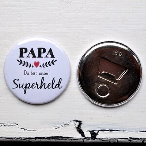 Könnte beinhalten: Ein weißer Knopf mit dem Wort "PAPA" über "Du bist unser Superheld" in schwarzer Schrift, mit einem roten Herz und Blättern. Daneben ein silberner Flaschenöffner mit der Zahl 59.