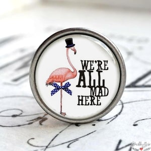 Könnte beinhalten: Ein weißer runder Knopf mit einem rosa Flamingo, der einen schwarzen Zylinder und eine blaue und weiße gepunktete Schleife trägt. Der Text "We're All Mad Here" ist in Schwarz auf dem Knopf gedruckt.