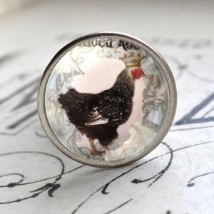 Könnte beinhalten: Ein silberner Knopf mit einer Glasoberfläche, die einen schwarz-weißen Hahn mit Krone zeigt. Der Hahn ist von einem weißen und grauen Blumendesign umgeben.