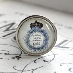 Könnte beinhalten: Ein silberner Knopf mit einer Glasoberfläche, die einen Kranz aus Lavendelblüten und eine Krone zeigt. Der Text "Les Fleurs de Lavande Savon" ist auf das Glas gedruckt.
