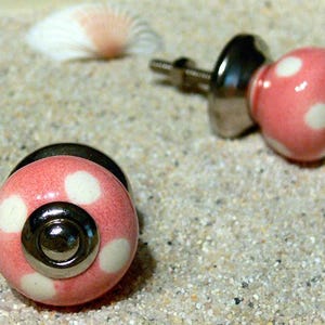 Könnte beinhalten: Zwei rosa Schrankknöpfe mit weißen Punkten und silbernen Beschlägen. Die Knöpfe sind rund und glänzend. Das Bild zeigt die Knöpfe und eine Muschel auf sandigem Untergrund. Ideal, um Möbeln eine verspielte Note zu verleihen.