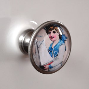 Könnte beinhalten: Ein silberner Knopf mit einem runden Glaseinsatz, der eine Vintage-Illustration eines Kindes in Matrosenkleidung zeigt, das ein Spielzeugsegelboot hält.