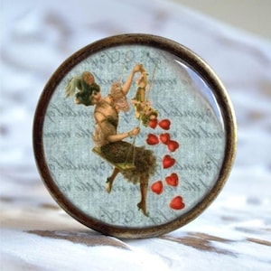 Könnte beinhalten: Ein runder, messingfarbener Knopf mit einem Glaseinsatz, der eine Vintage-Illustration einer Frau auf einer Schaukel zeigt, die rote Herzen verstreut. Der Hintergrund ist ein blaues und weißes gemustertes Papier mit kursivem Schriftzug.