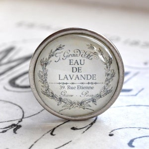 Könnte beinhalten: Ein runder, silberfarbener Schubladengriff mit Glaseinsatz. Der Einsatz zeigt den Text "Eau de Lavande" in einem dekorativen Kranz, zusammen mit der Adresse "39. Rue Etienne, Grasse - Paris."