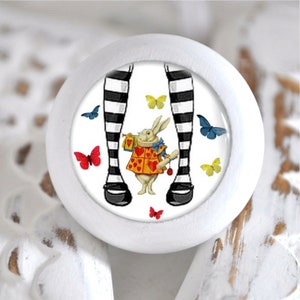 Könnte beinhalten: Weißer, runder Schrankknopf mit einer skurrilen Illustration. Das Design zeigt ein Kaninchen in einer Weste, schwarz-weiß gestreifte Beine und bunte Schmetterlinge. Ein dekoratives Element für Möbel.