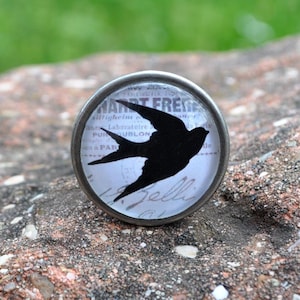 Könnte beinhalten: Ein silberner Knopf mit einer schwarzen Silhouette eines Vogels auf weißem Hintergrund mit dem sichtbaren Text "MART FRE" und "BELL".