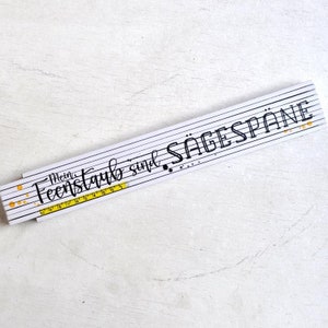 Könnte beinhalten: Ein weißer Zollstock mit schwarzen Linien und dem Text "Mein Feenstaub sind Sägespäne". Der Zollstock hat eine gelbe Maßeinteilung in Zoll. Goldfarbene Farbspritzer und schwarze Punkte akzentuieren das Design.