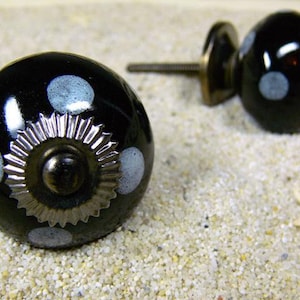 Könnte beinhalten: Zwei schwarze Keramik-Schrankknöpfe mit weißen Tupfen. Ein Knopf hat eine dekorative silberfarbene Metallbasis. Die Knöpfe liegen auf einer sandigen Oberfläche, mit einem Seestern im Hintergrund. Diese dekorativen Knöpfe sind perfekt, um Möbeln eine einzigartige Note zu verleihen.