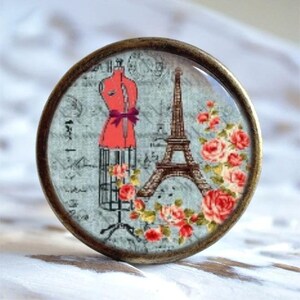 Könnte beinhalten: Ein runder, messingfarbener Knopf mit einem Glaseinsatz, der ein Vintage-Pariser-Design zeigt. Das Design umfasst eine rote Schneiderpuppe, den Eiffelturm und rosafarbene Rosen.