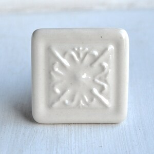 Könnte beinhalten: Quadratischer, cremefarbener Keramik-Schrankknopf mit erhabenem, symmetrischem Blumendesign in der Mitte. Der Knopf hat abgerundete Kanten und eine glänzende Oberfläche, geeignet für Möbel oder Dekorationszwecke.