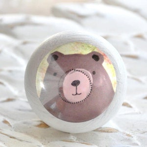 Könnte beinhalten: Weißer Holz-Knopf mit einem runden, bunten Aufkleber, der einen braunen Cartoon-Bären mit rosa Nase und schwarzen Augen zeigt.