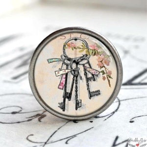 Könnte beinhalten: Ein silberner Knopf mit einer Glasoberfläche, die eine Vintage-Illustration von Schlüsseln zeigt, die mit bunten Bändern und einem Blumenzweig zusammengebunden sind. Die Schlüssel sind mit einer Schleife zusammengebunden und der Text "STC" ist in der oberen linken Ecke des Bildes sichtbar.