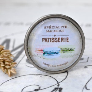 Könnte beinhalten: Ein silberner Knopf mit einem weißen und blauen Makronen-Design. Der Knopf trägt den Text "Spécialité Macarons Patisserie Marque de Fabrique".