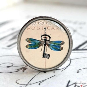 Könnte beinhalten: Ein runder, silberfarbener Knopf mit einem Libellen- und Schlüsseldesign. Die Libelle hat blau-grüne Flügel, und der Schlüssel ist schwarz. Der Hintergrund zeigt die Worte "POST CARD".