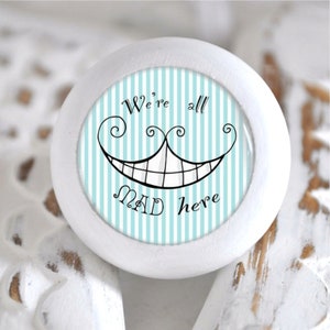 Könnte beinhalten: Runder, weißer Möbelknopf mit blau-weiß gestreiftem Mittelteil. In der Mitte befindet sich ein lächelndes Gesicht der Grinsekatze mit dem Text "We're all MAD here" in schwarzer Schrift. Der Knopf ist wahrscheinlich für eine Schublade oder einen Schrank.