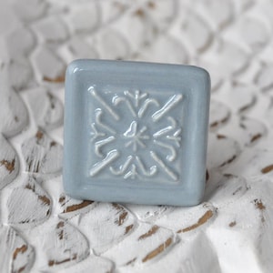 Könnte beinhalten: Ein quadratischer Keramik-Knopf mit einem erhabenen, weißen, geometrischen Design. Der Knopf ist hellblau.