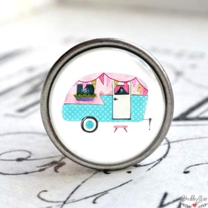Könnte beinhalten: Ein weißer Keramik-Knopf mit einem silbernen Metallring. Der Knopf zeigt einen rosa-blauen Wohnwagen mit einer weißen Tür und einem kleinen rosa Hocker davor. Der Wohnwagen hat ein blaues und weißes Punkte-Muster und eine rosa Girlande.