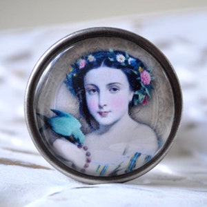 Könnte beinhalten: Ein runder Anhänger in Silberton mit einer Glaskuppel, die eine Vintage-Illustration einer Frau mit einer Blumenkrone und einem blauen Vogel zeigt, der auf ihrer Schulter sitzt.