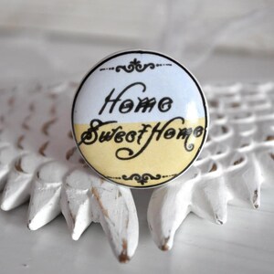 Könnte beinhalten: Weißer und gelber Keramik-Knopf mit schwarzer Aufschrift "Home Sweet Home".