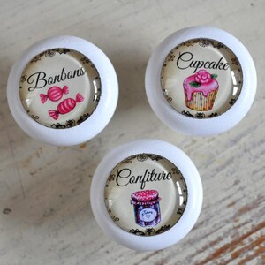 Könnte beinhalten: Drei weiße runde Knöpfe mit Glaseinsätzen. Jeder Knopf hat ein anderes Design: einer mit Süßigkeiten, einer mit einem Cupcake und einer mit einem Marmeladentopf. Der Text auf den Knöpfen lautet "Bonbons", "Cupcake" und "Confiture".