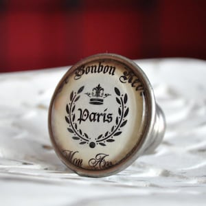 Könnte beinhalten: Ein silberfarbener Flaschenverschluss mit einer weißen runden Oberseite, die mit einer schwarzen Krone, einem Kranz und dem Text "Bonbon Kic", "Paris" und "Mon Amie" verziert ist.