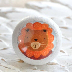 Könnte beinhalten: Weißer Holz-Knopf mit einem runden, bunten Aufkleber eines Cartoon-Löwen mit orangefarbenem Fell und einer roten Mähne. Der Löwe hat braune Augen und eine rosa Nase.