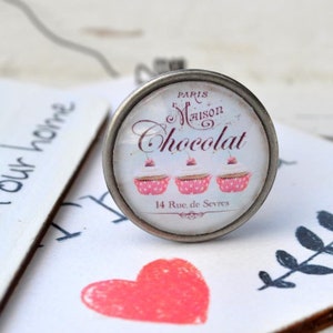 Könnte beinhalten: Ein silberner Knopf mit einem Glaseinsatz, der ein Vintage-Design einer Pariser Bäckerei zeigt. Das Design enthält den Text "Paris Maison Chocolat" und drei rosafarbene Cupcakes mit Kirschen obenauf. Der Text "14 Rue de Sevres" ist ebenfalls enthalten.