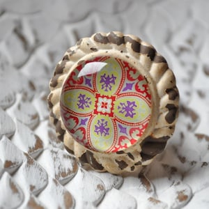 Könnte beinhalten: Ein dekorativer Schrankknopf mit einem gemusterten Glaseinsatz. Der Knopf hat eine cremefarbene, strukturierte Basis mit braunen Akzenten. Der Glaseinsatz zeigt ein rotes, gelbes und violettes Blumenmuster. Geeignet für Möbel und Heimdekoration.