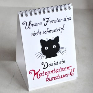 May include: A white desk calendar with a black cat silhouette and the text "Unsere Fenster sind nicht schmutzig! Das ist ein Katzentatzen-kunstwerk!"