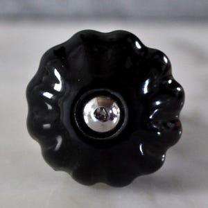 Könnte beinhalten: Ein glänzender schwarzer Schrankknopf mit gewelltem Rand und silberfarbener Mitte. Der Knopf hat eine blütenartige Form und besteht wahrscheinlich aus Keramik oder einem ähnlichen Material. Das Design ist schlicht und elegant.