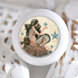 Könnte beinhalten: Weißer runder Knopf mit einer Meerjungfrauen-Illustration. Die Meerjungfrau hat langes schwarzes Haar und einen blauen Schwanz. Sie sitzt auf einem Felsen mit blauen Seesternen um sie herum.