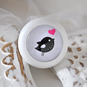 Könnte beinhalten: Weißer runder Knopf mit einem schwarz-weißen Vogelmotiv und einem rosa Herzen. Der Vogel schaut nach rechts und hat einen weißen Bauch und schwarze Punkte. Das Herz befindet sich über dem Kopf des Vogels.