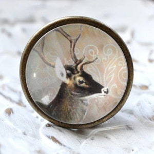 Könnte beinhalten: Ein runder, bronzefarbener Knopf mit einem Glaseinsatz, der ein detailliertes Bild eines Hirsches mit Geweih vor einem hellbraunen Hintergrund mit einem weißen Blumenmuster zeigt.