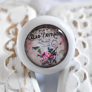 Könnte beinhalten: Weißer Keramik-Knopf mit einem runden, bunten Bild einer Vintage-Kakaowerbung mit einer Teekanne, Blumen und dem Text "Tenez à votre déjeuner Le Cacao-Payraud".