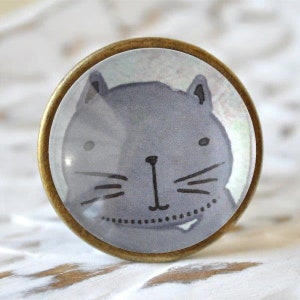 Könnte beinhalten: Ein runder, messingfarbener Knopf mit einem Glaseinsatz, der eine graue Comic-Katze mit schwarzen Schnurrhaaren und Augen zeigt. Die Katze hat einen kleinen, weißen Zahn, der herausragt.