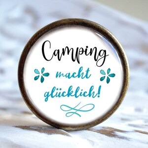 Könnte beinhalten: Ein runder, weißer Knopf mit einem braunen Metallrahmen. Der Knopf hat blaue Aquarellblumen und den Text "Camping macht glücklich!" in Schwarz.