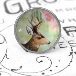 Könnte beinhalten: Ein silberfarbener Knopf mit einem Glascabochon, der eine Vintage-Illustration eines Hirsches mit Geweih und einer rosa Rose zeigt. Der Hintergrund ist in einem sanften Grün gehalten.