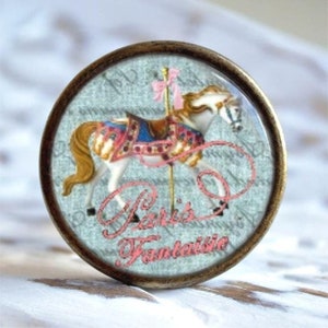 Könnte beinhalten: Ein runder, messingfarbener Knopf mit einem Glaseinsatz, der eine Vintage-Illustration eines weißen Karussells mit einem rosafarbenen Sattel und rosafarbenen Bändern zeigt. Der Hintergrund ist hellblau mit einem verblassten, handschriftlichen Textmuster. Der Text "Paris Fantasie" ist in rosa Kursivschrift geschrieben.