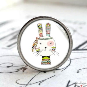 Könnte beinhalten: Ein weißes Kaninchen mit bunten Tribal-Mustern an Ohren, Gesicht und Hals. Das Kaninchen trägt ein Stirnband mit einer Feder. Das Bild ist auf einem runden, silbernen Knopf.