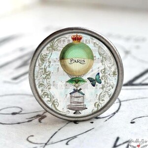 Könnte beinhalten: Ein silberner Knopf mit einer Glasoberfläche, die eine Vintage-Illustration eines Heißluftballons mit dem Wort "PARIS" zeigt. Der Ballon ist grün und gelb mit einer Krone oben. Ein blauer Schmetterling fliegt neben dem Ballon. Der Hintergrund ist ein weißes und graues Muster.