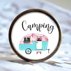 Könnte beinhalten: Ein runder, weißer Keramik-Knopf mit einem braunen Metallring am Rand. Der Knopf zeigt einen rosafarbenen und blauen Wohnwagen mit weißen Punkten und dem Wort "Camping" in schwarzer Kursivschrift.