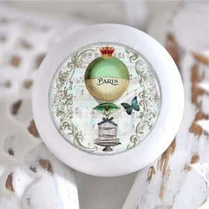 Könnte beinhalten: Weißer runder Knopf mit einem Vintage-Design, das einen Heißluftballon mit dem Wort "PARIS" darauf, einen Vogelkäfig und einen Schmetterling zeigt. Der Hintergrund ist hellblau mit Noten und einem dekorativen Rand.