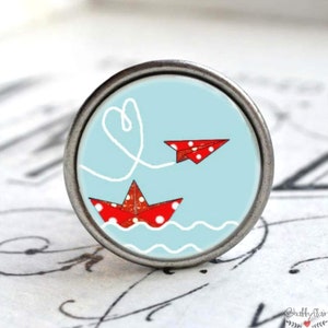 Könnte beinhalten: Ein runder, silberner Knopf mit blauem Hintergrund und einem roten Papierboot und einem Flugzeug mit weißen Punkten. Das Flugzeug fliegt vom Boot weg, verbunden durch eine weiße Schnur, die eine Herzform bildet.