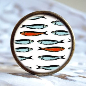 Könnte beinhalten: Ein runder, messingfarbener Knopf mit weißem Hintergrund und einem Muster aus blauen und orangefarbenen Fischen. Die Fische sind in Reihen angeordnet und zeigen in verschiedene Richtungen.