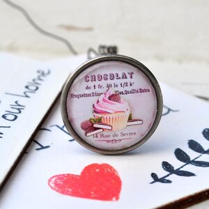 Könnte beinhalten: Ein silberner Ring mit einem runden Glascabochon, der eine Vintage-Werbung für Schokoladen-Cupcakes zeigt. Der Text lautet "CHOCOLAT de 1 fr. 40 le 1/2 k Croquettes Bâton les, Qualité Extra 14 Rue de Sevres".